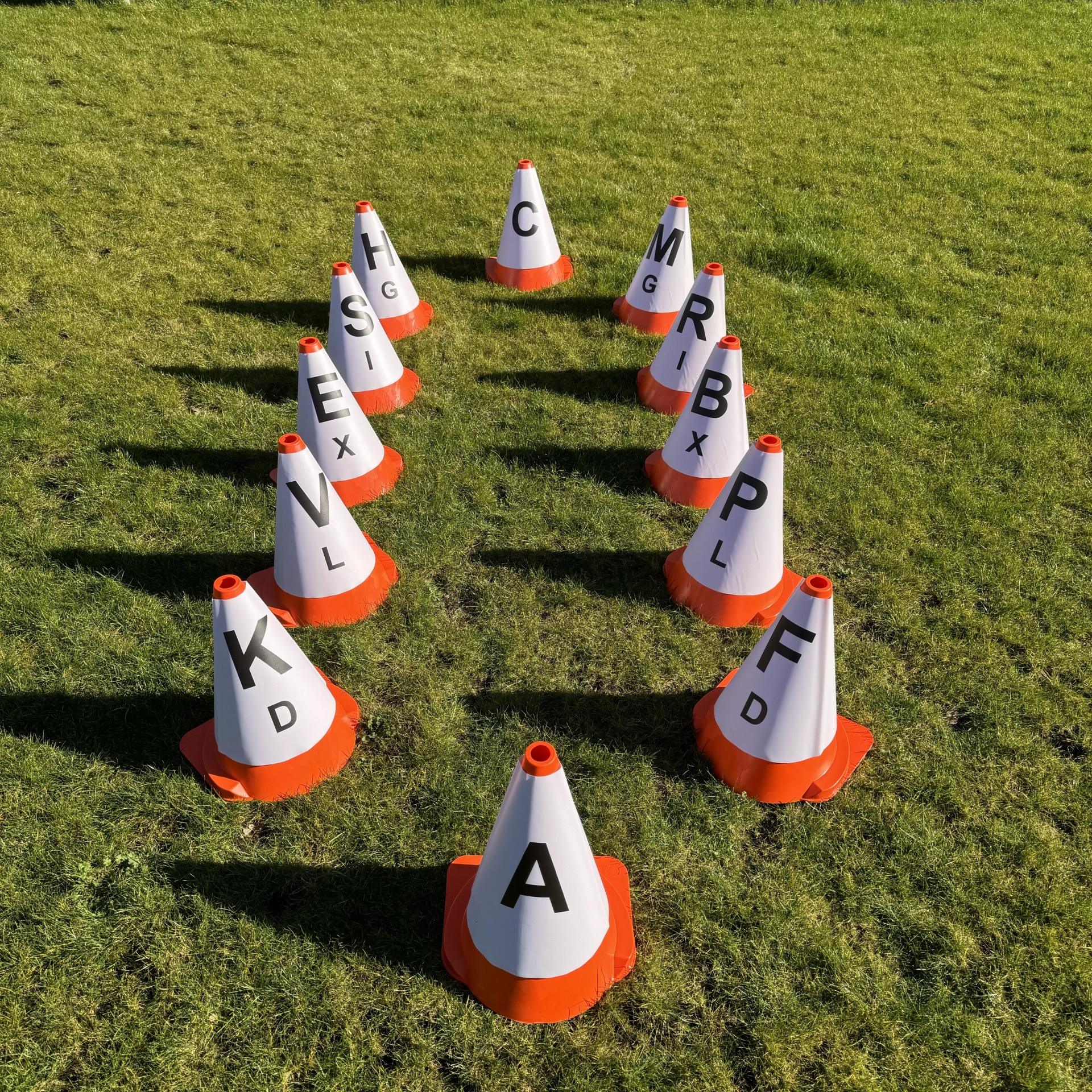 Doze cones de trânsito brancos com base alaranjada e letras pretas sobre relvado verde formando uma seta.