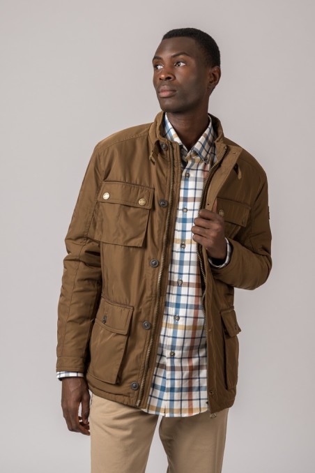 Homem com casaco castanho, camisa xadrez e calças bege em fundo cinzento claro