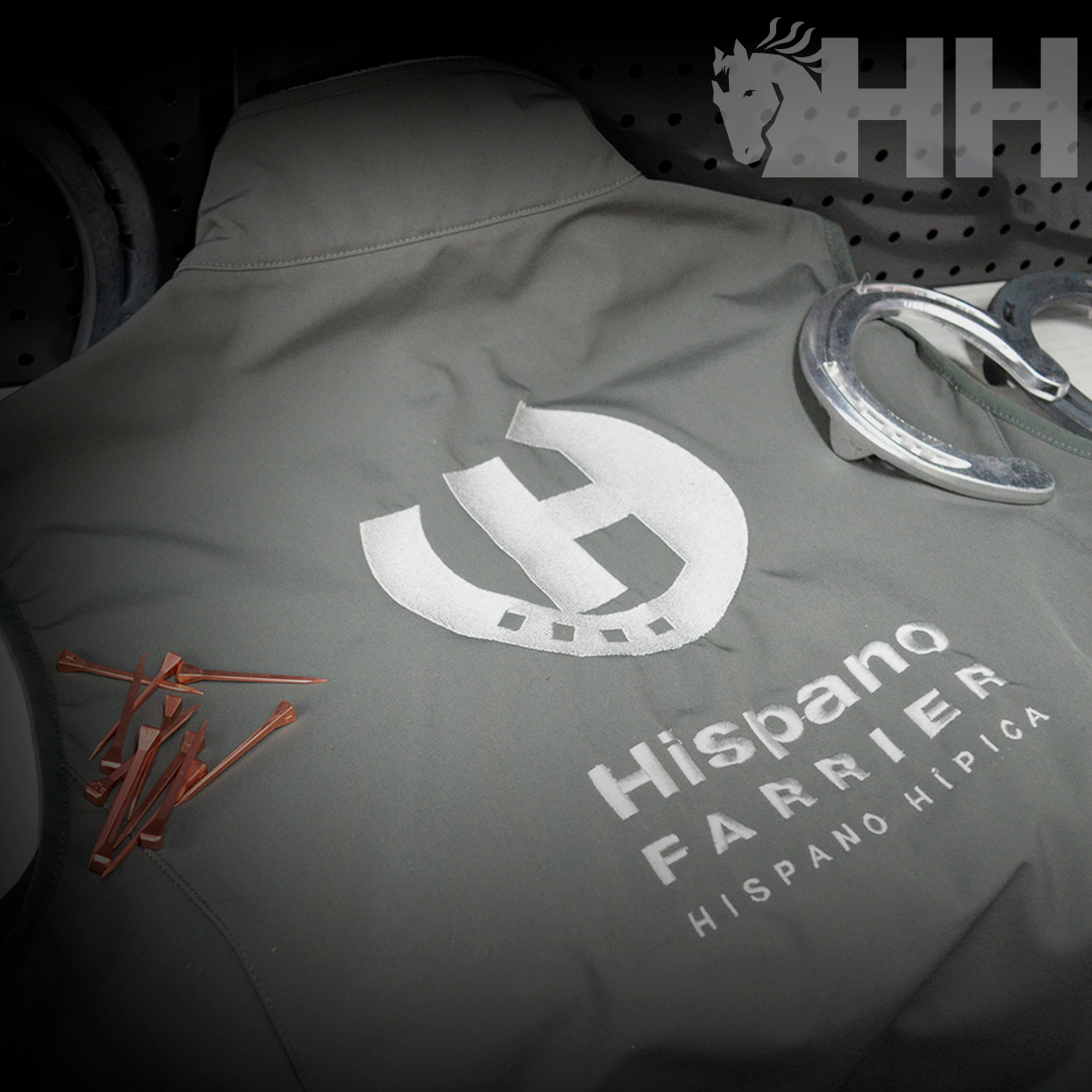 Casaco cinza com logótipo e texto Hispano Farrier, com ferradura prateada e pregos em fundo metálico.
