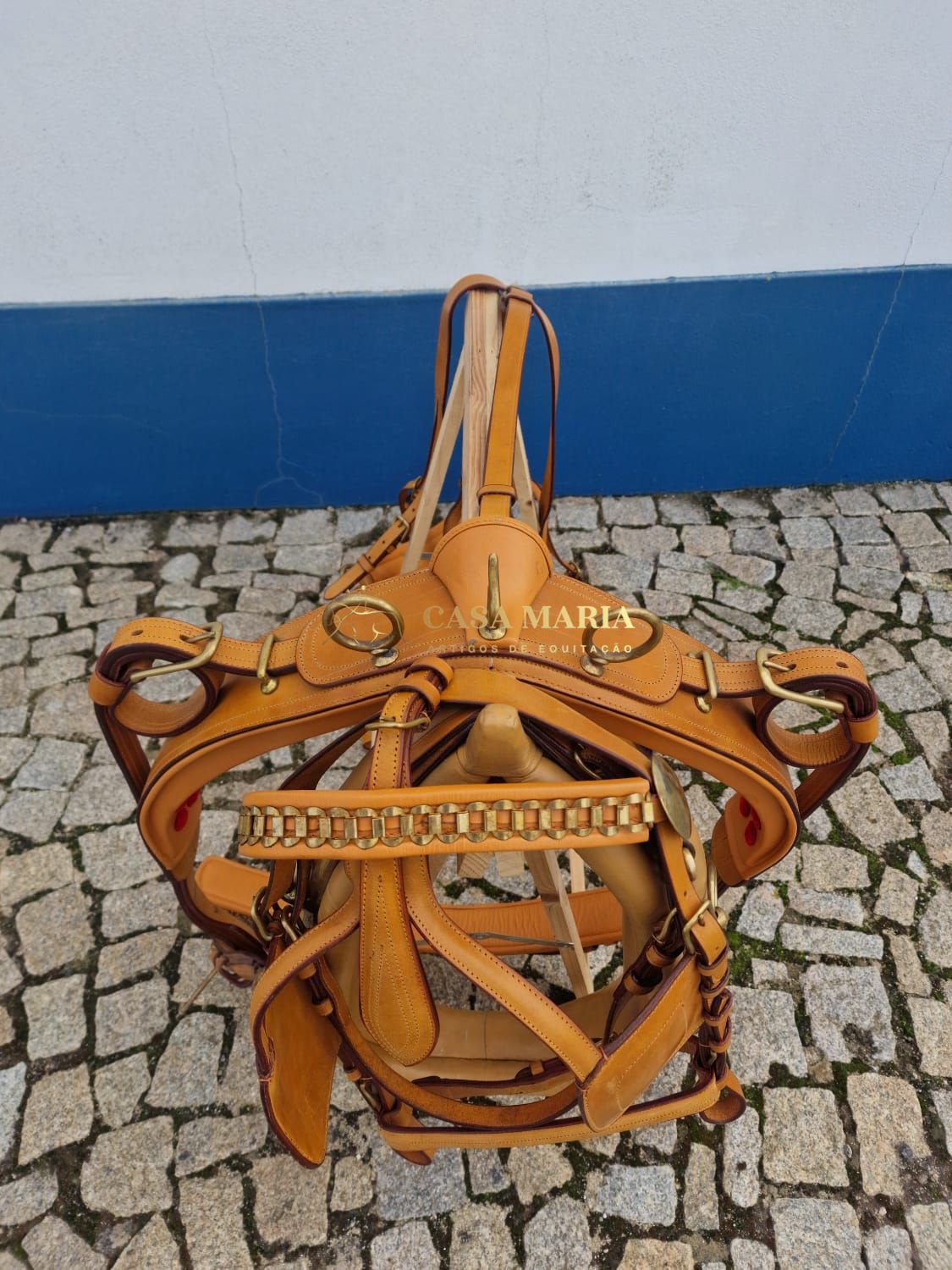Peitoral para cavalo em couro castanho com fivelas douradas numa calçada portuguesa.