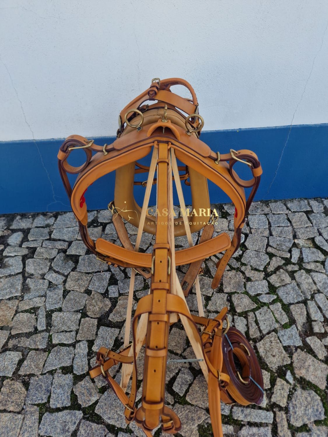 Sela de equitação em couro castanho com detalhes metálicos sobre chão de pedra e parede branca com faixa azul