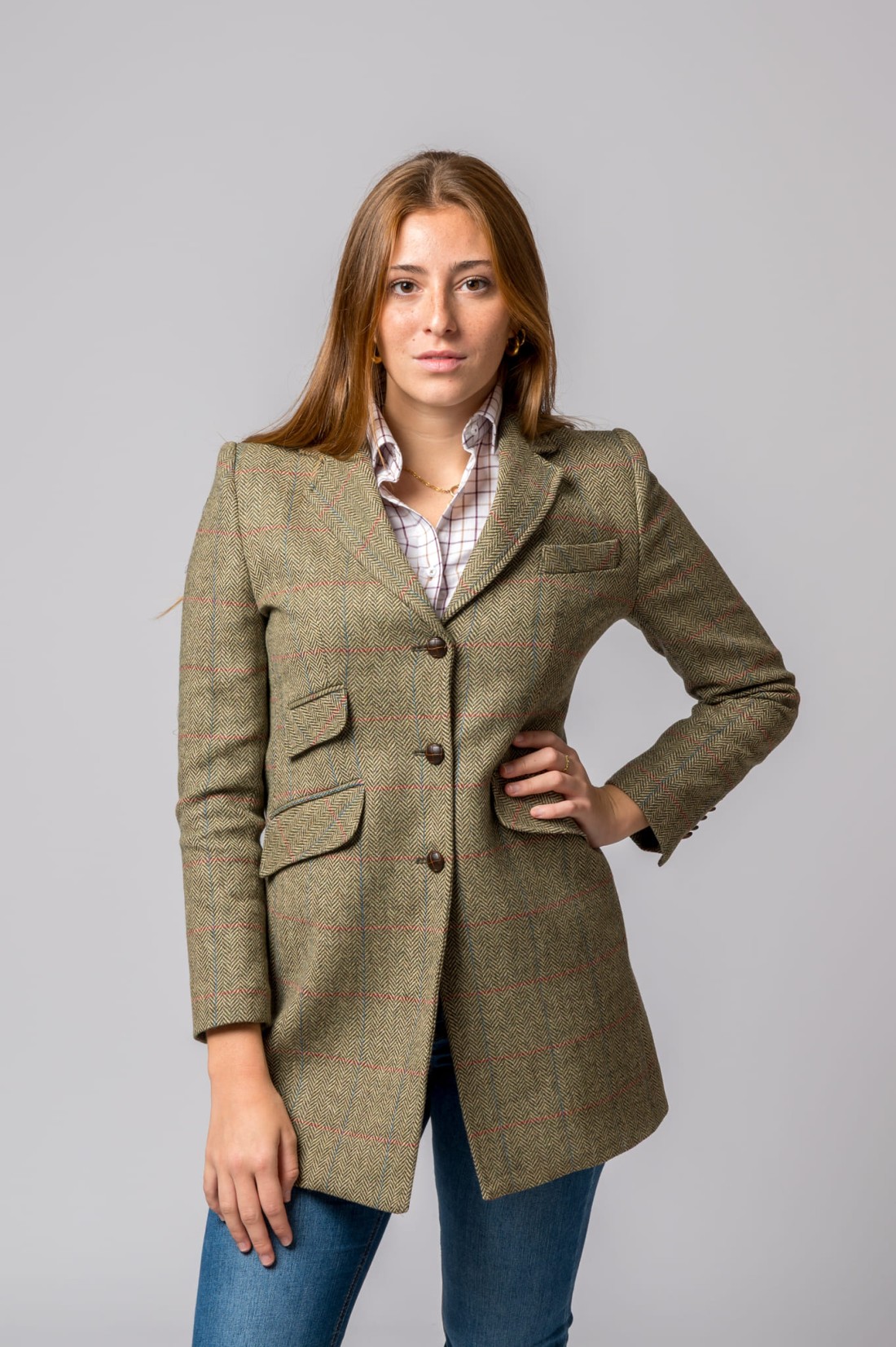 Mulher de blazer verde castanho com padrão espinha de peixe e camisa branca xadrez em fundo neutro