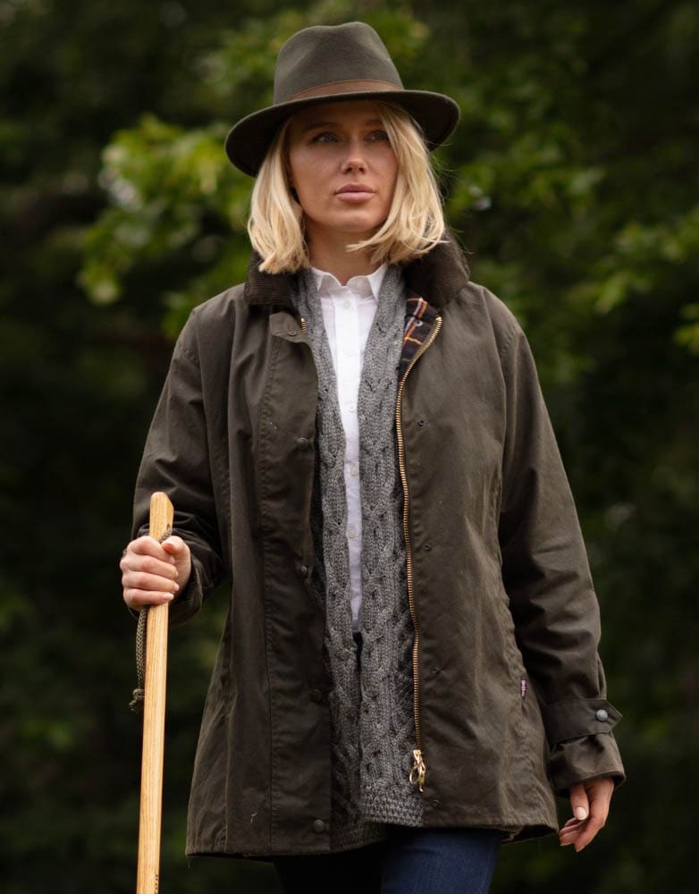 Mulher vestindo casaco verde escuro, cardigan cinzento e chapéu castanho em ambiente exterior