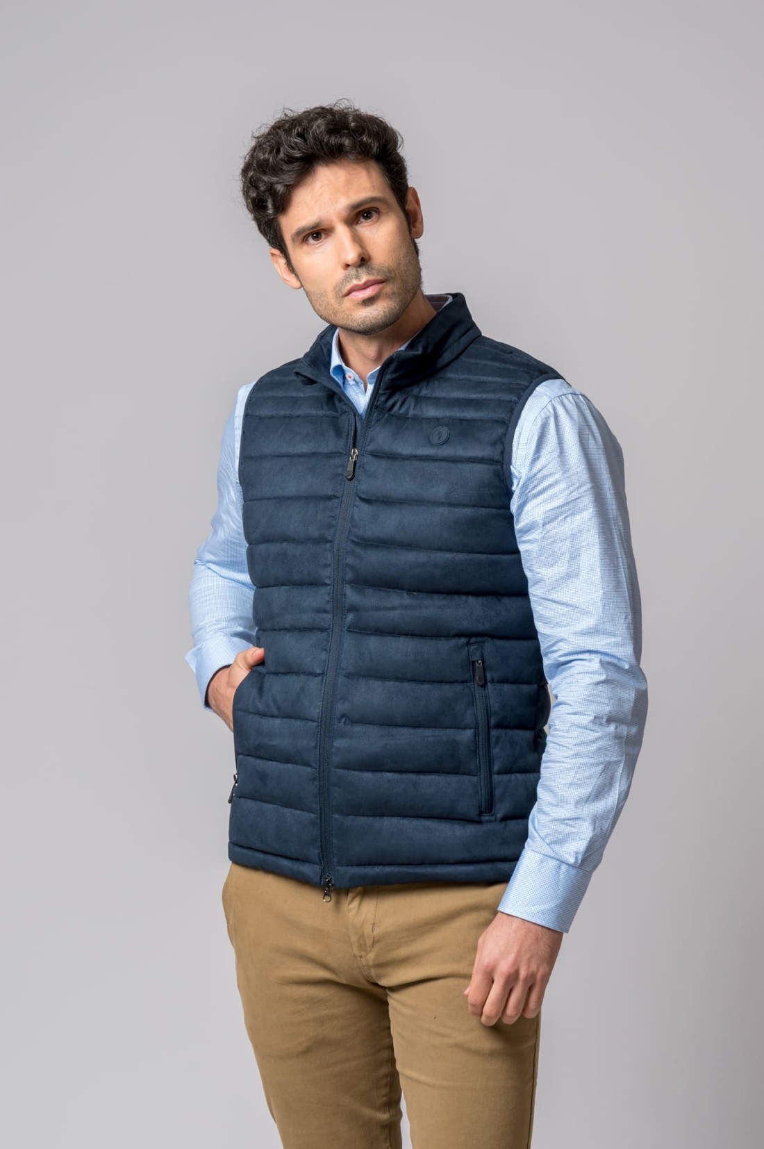 Colete azul escuro acolchoado com fecho de correr, usado por homem com camisa azul clara e calças castanhas.