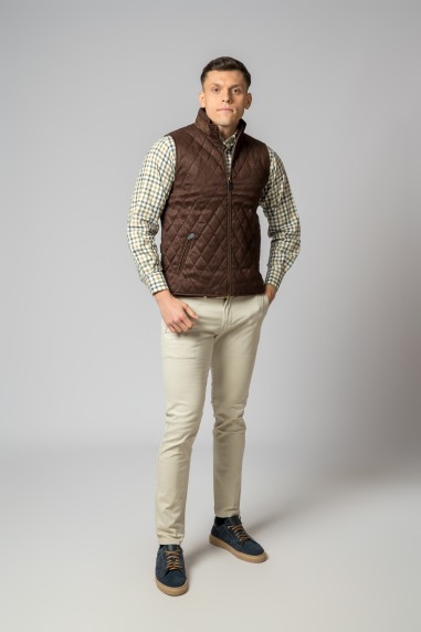 Homem com colete castanho, camisa xadrez, calças bege e sapatos escuros em fundo cinza