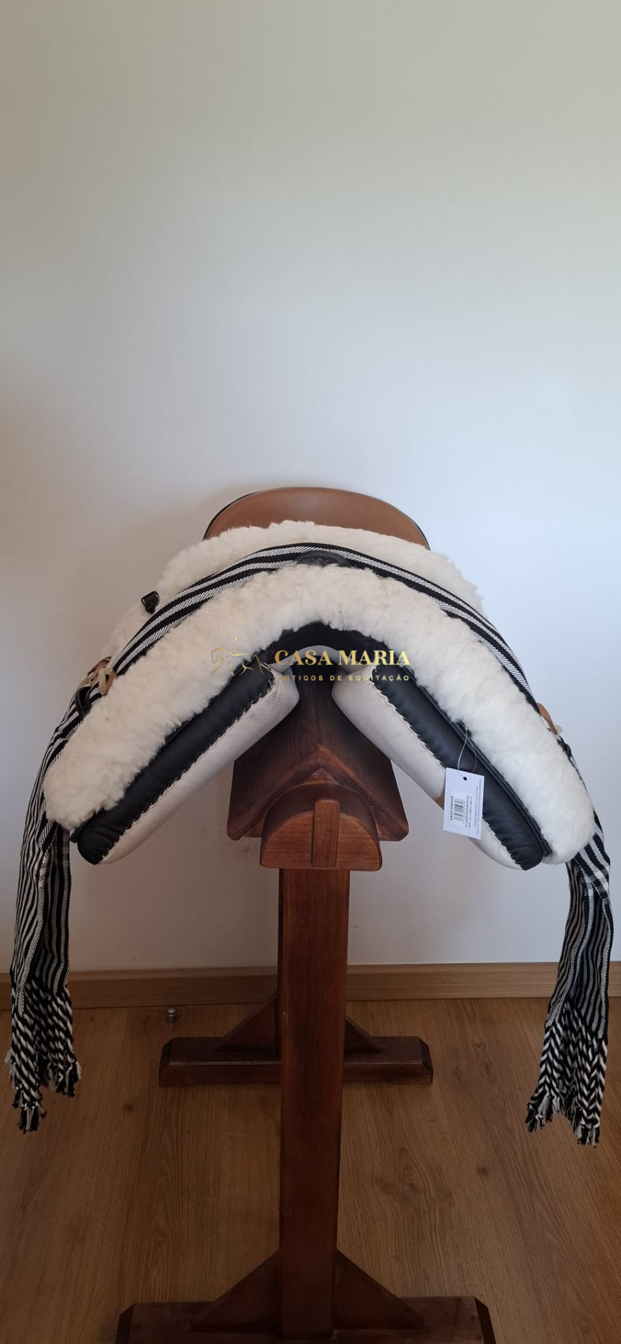Persiana de equitação com pele branca, couro preto e tecido riscado a preto e branco num suporte de madeira.
