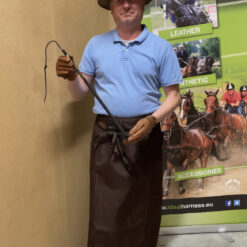 Homem com chapéu, camisa azul clara, avental e luvas castanhos a segurar chicote em frente a cartaz promocional