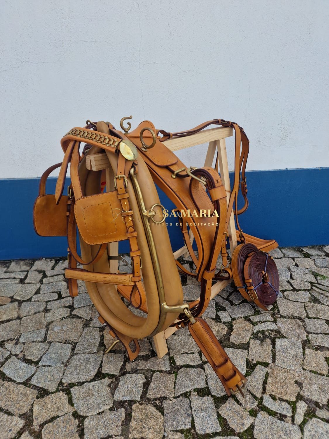 Arreio de couro castanho claro com acessórios pendurado em suporte de madeira sobre chão de pedras