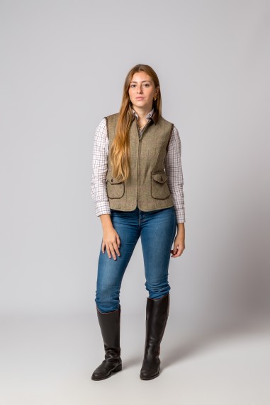 Mulher com colete tweed castanho, camisa xadrez branca, jeans azul e botas pretas em fundo cinzento