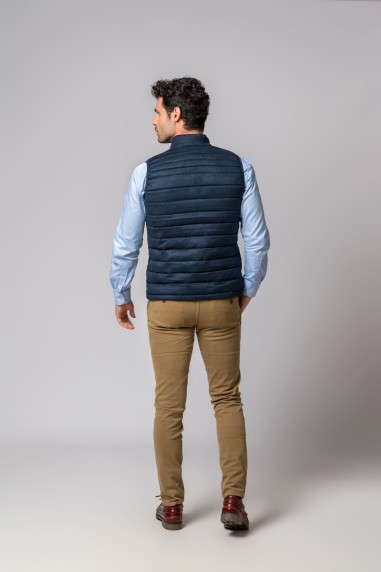 Homem de costas com colete azul acolchoado, camisa azul clara, calças castanhas e sapatos castanhos