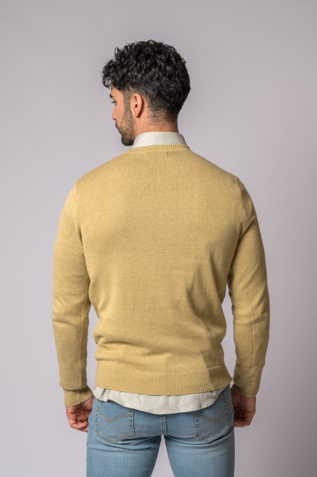 Homem visto de costas com camisola bege e camisa cinza clara por baixo, veste jeans azul claro, fundo cinzento