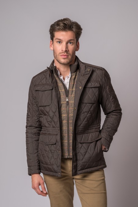Homem a usar casaco acolchoado castanho escuro com bolsos e camisa branca num fundo cinzento.