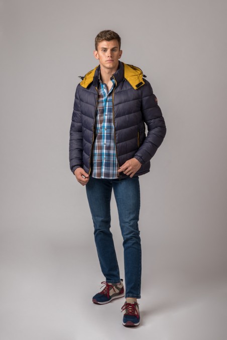 Homem veste casaco acolchoado azul escuro com capuz amarelo, camisa xadrez azul, jeans e ténis coloridos.