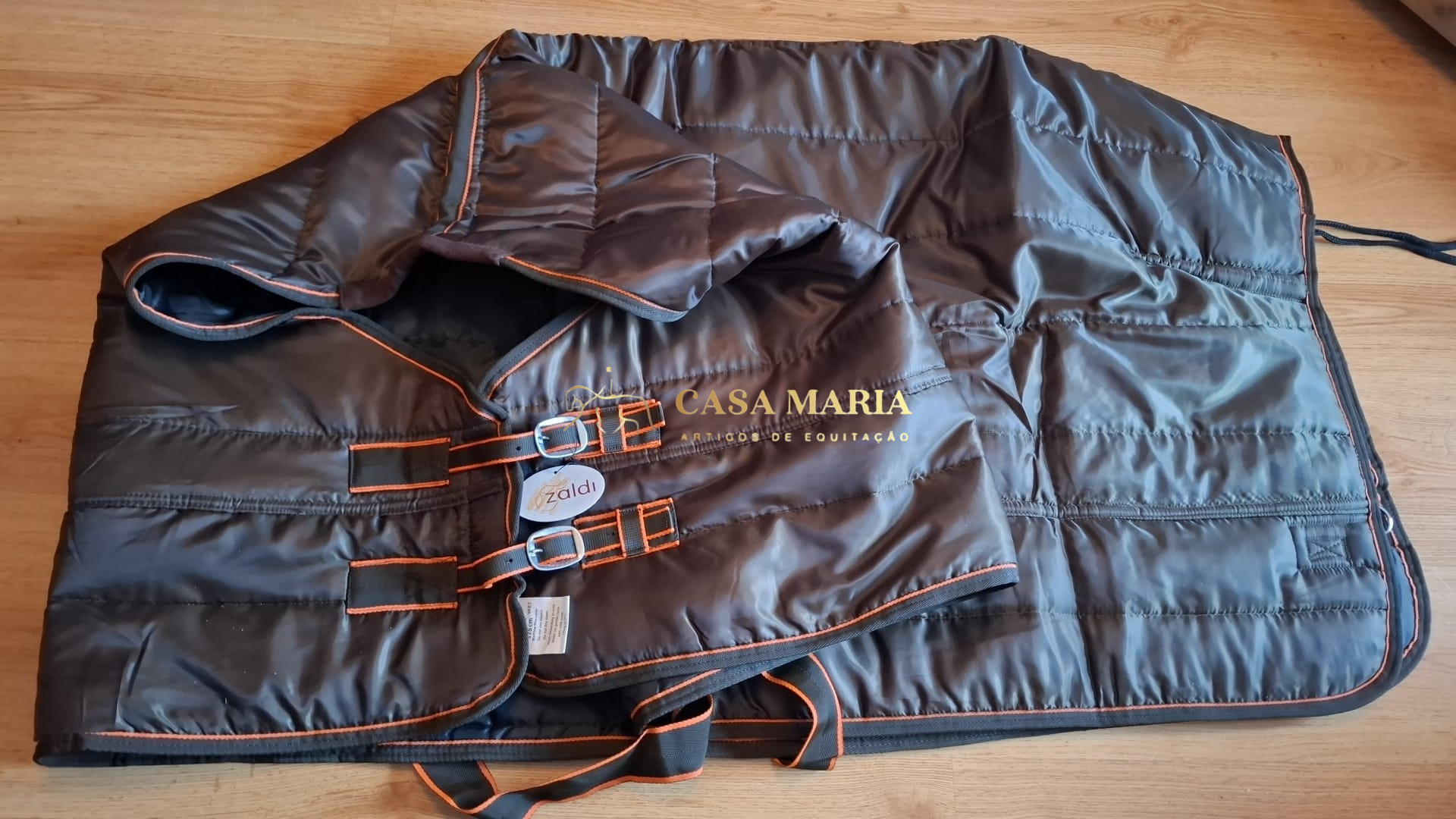 Manta acolchoada de equitação preta com costuras laranja e fivelas, sobre piso amadeirado
