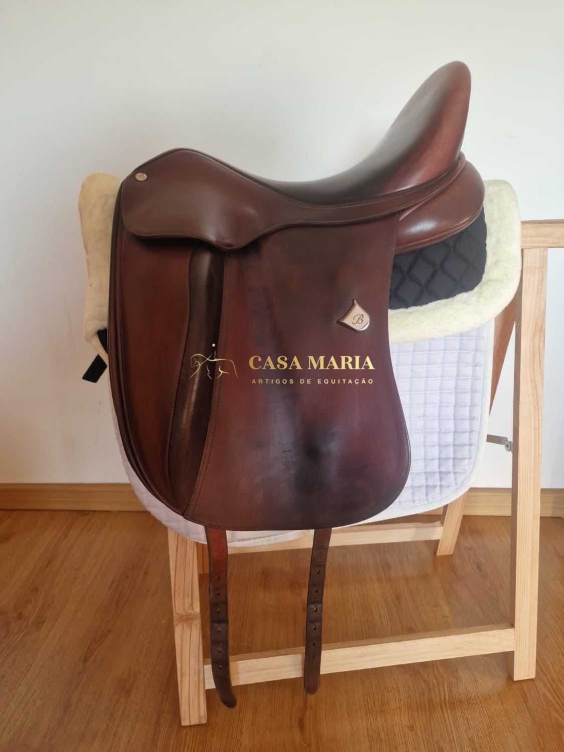 Sela de cavalo de couro castanho pendurada em suporte de madeira com manta acolchoada branca e negra