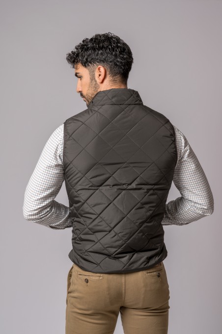 Homem visto de costas usando colete acolchoado preto com padrão losango, camisa branca xadrez e calças castanhas.