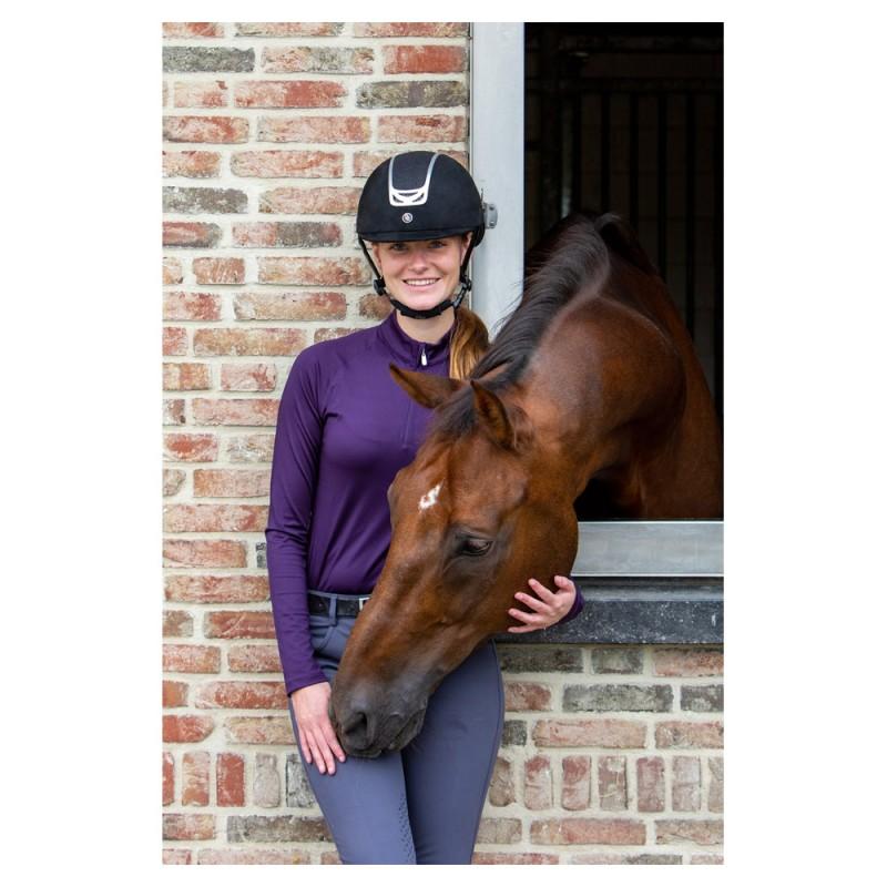 Mulher com capacete e roupa de equitação abraça cavalo castanho ao lado de muro de tijolos.