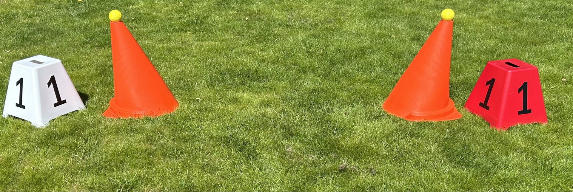 Cones laranjas com bolas amarelas no topo e cubos numerados 11, branco e vermelho, sobre relva.