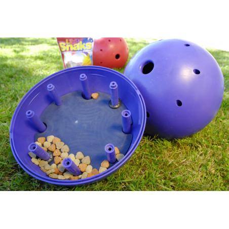 Dois dispensadores de comida para cães em forma de bola, um aberto com petiscos visíveis e outro fechado, embalagem 'Shaks' ao fundo sobre relva