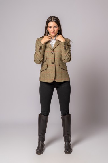 Mulher com blazer bege de risca de giz, camisa xadrez, calças pretas e botas castanho-escuro em fundo cinza.