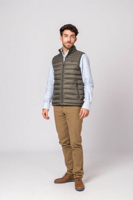 Homem vestindo colete acolchoado verde oliva, camisa azul clara listrada, calças castanhas e sapatos castanhos