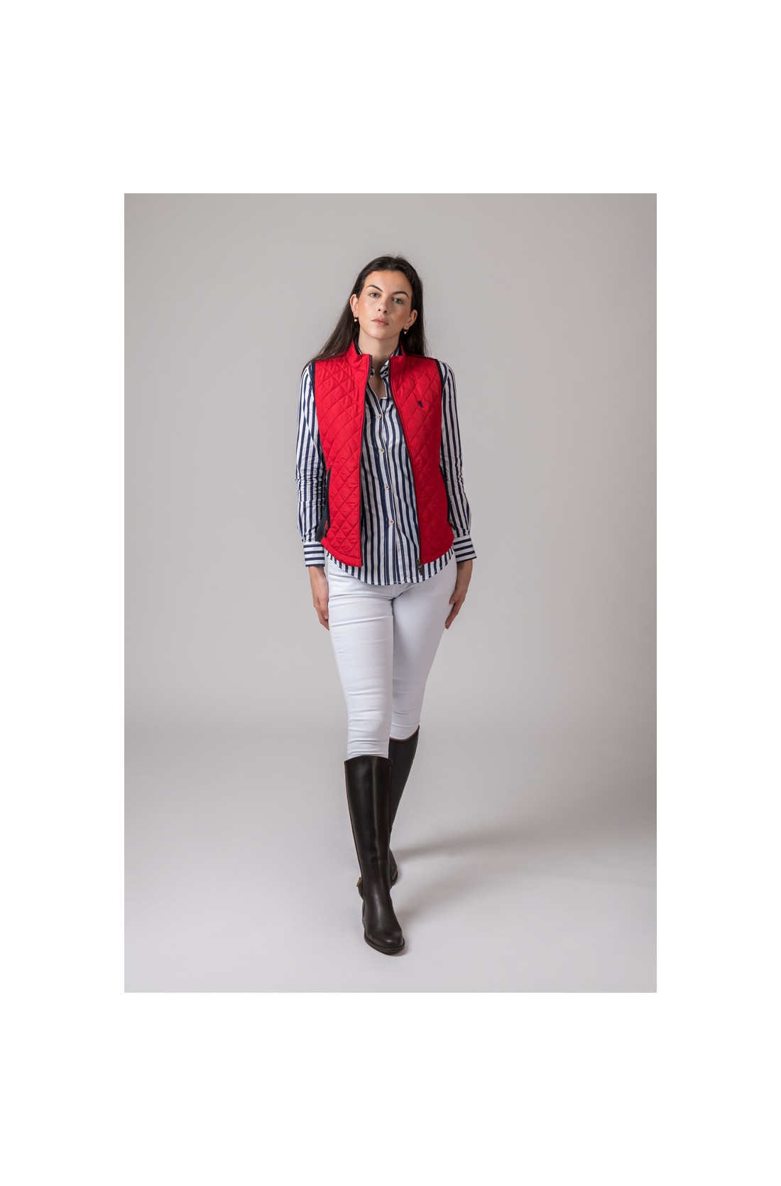 Mulher vestida com blusa listrada azul e branca, colete vermelho acolchoado e calças brancas com botas pretas