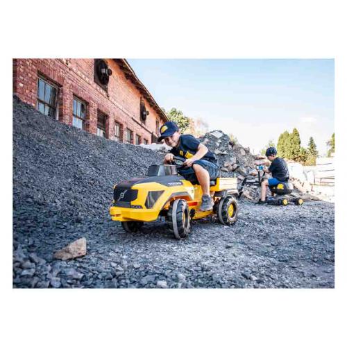 Crianças a andar em carrinhos de pedal amarelos em terreno pedregoso junto a um edifício de tijolos