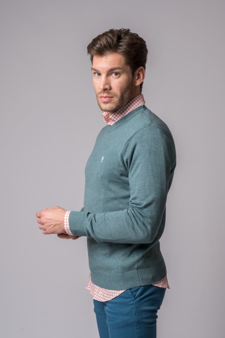 Homem com camisola azul esverdeada e camisa xadrez cor-de-rosa em fundo cinzento