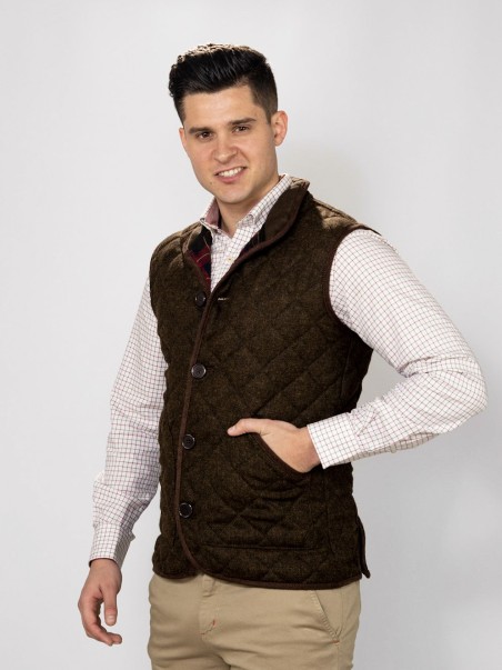Colete acolchoado castanho em padrão xadrez com botão e bolso, usado sobre camisa branca e calças bege