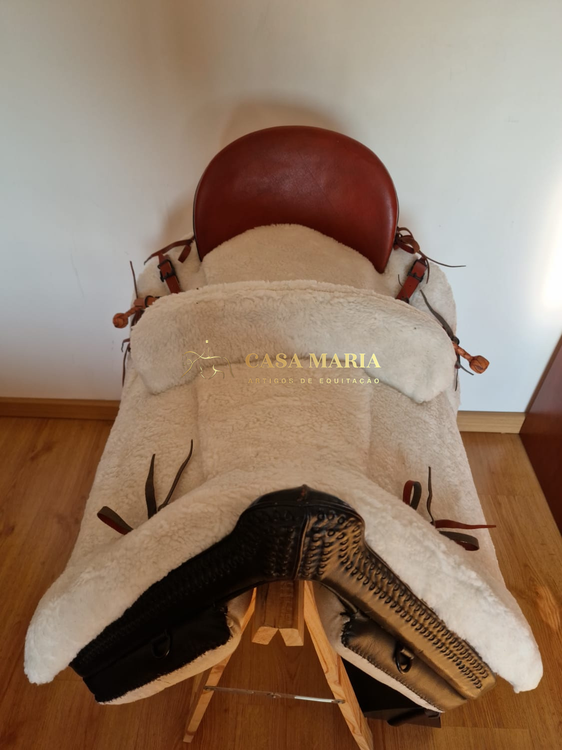 Sela de equitação em couro castanho e pelúcia branca sobre suporte de madeira