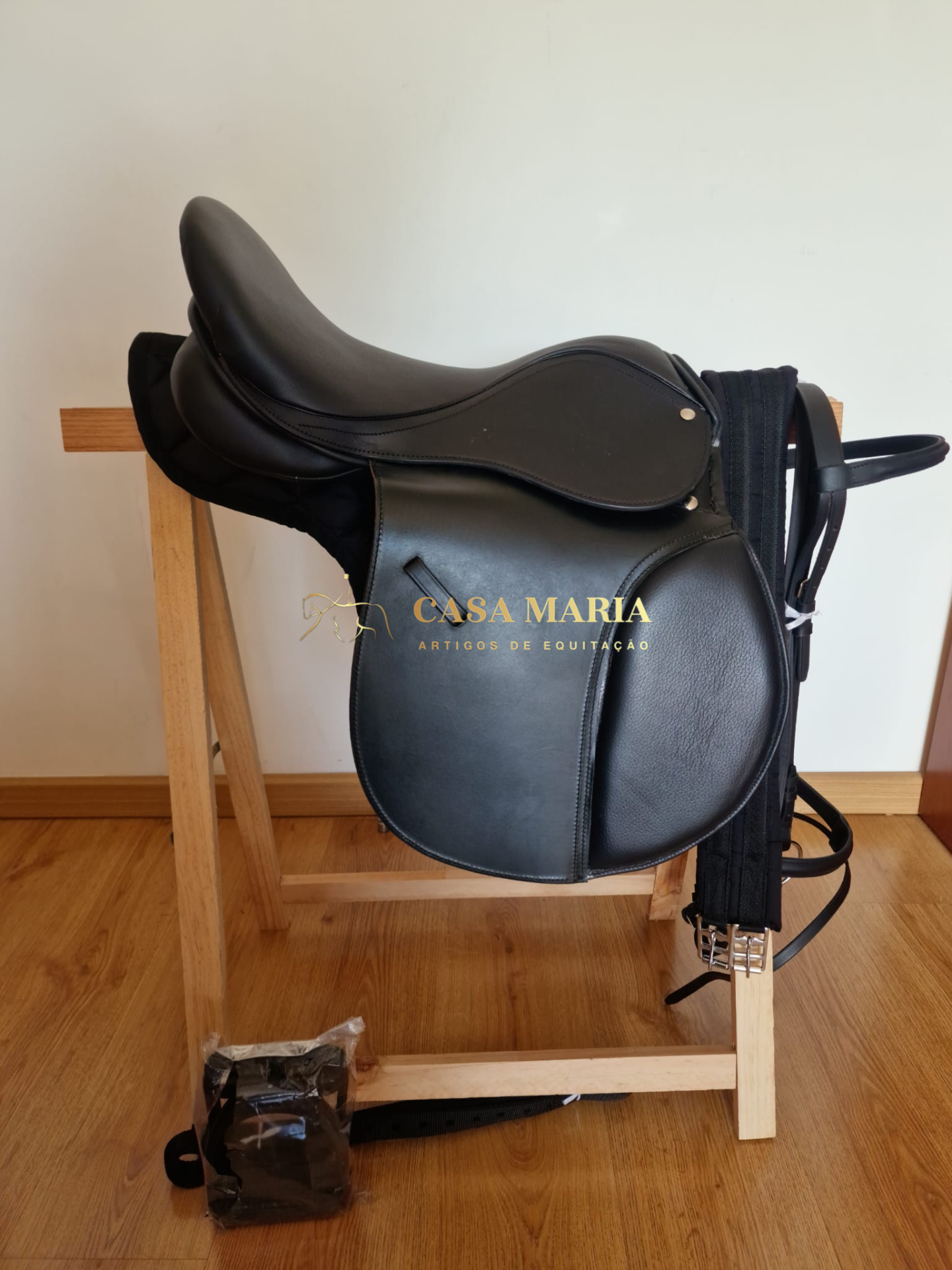 Sela de equitação preta em couro numa estrutura de madeira clara, com acessórios pendurados e embalagem no chão