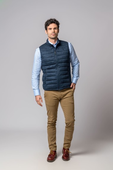 Homem com colete azul escuro acolchoado, camisa azul clara, calças castanhas e sapatos castanhos vermelhos.