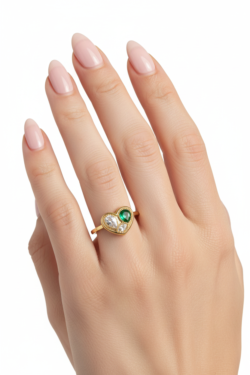 Anel dourado com pedras verde e brancas em forma de coração numa mão com unhas pintadas de rosa claro.