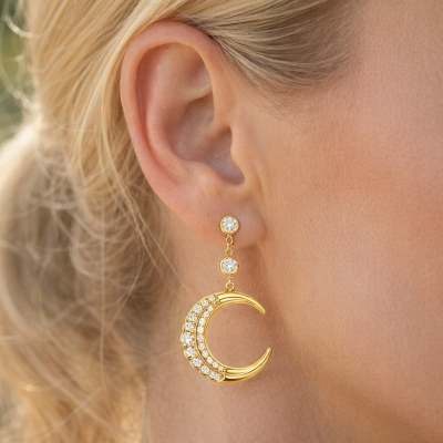Brinco pendente dourado com pedras brancas em forma de lua crescente na orelha de mulher.