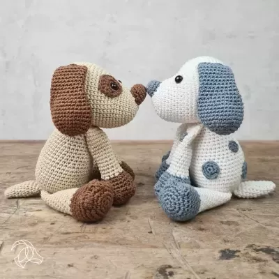 Dois cães de peluche em croché, um bege castanho e outro branco azul, sentados face a face sobre madeira