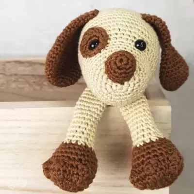 Boneco de crochê em forma de cão com corpo bege e detalhes castanhos apoiado numa caixa de madeira