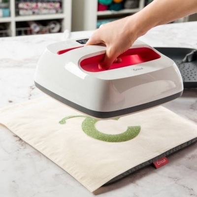 Ferro de engomar portátil branco e vermelho sobre saco de tecido bege com letra C verde numa bancada de mármore