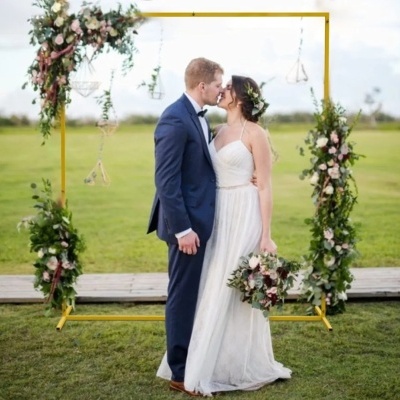 Casal vestido para casamento junto a arco decorado com flores e vegetação no exterior