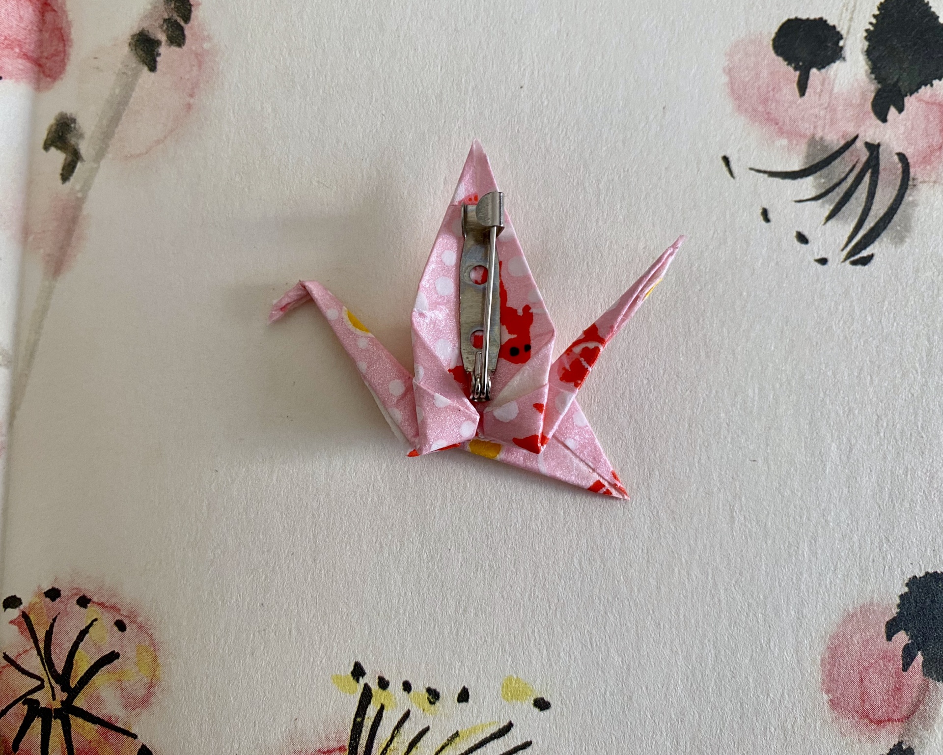 Pregadeira Tsuru Broche em forma de grua em papel rosa com fecho metálico sobre fundo florido.