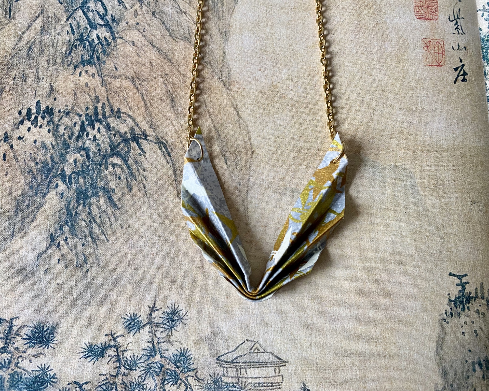 Colar Niki Colar com pendente de papel dobrado em correntes douradas sobre fundo com arte oriental