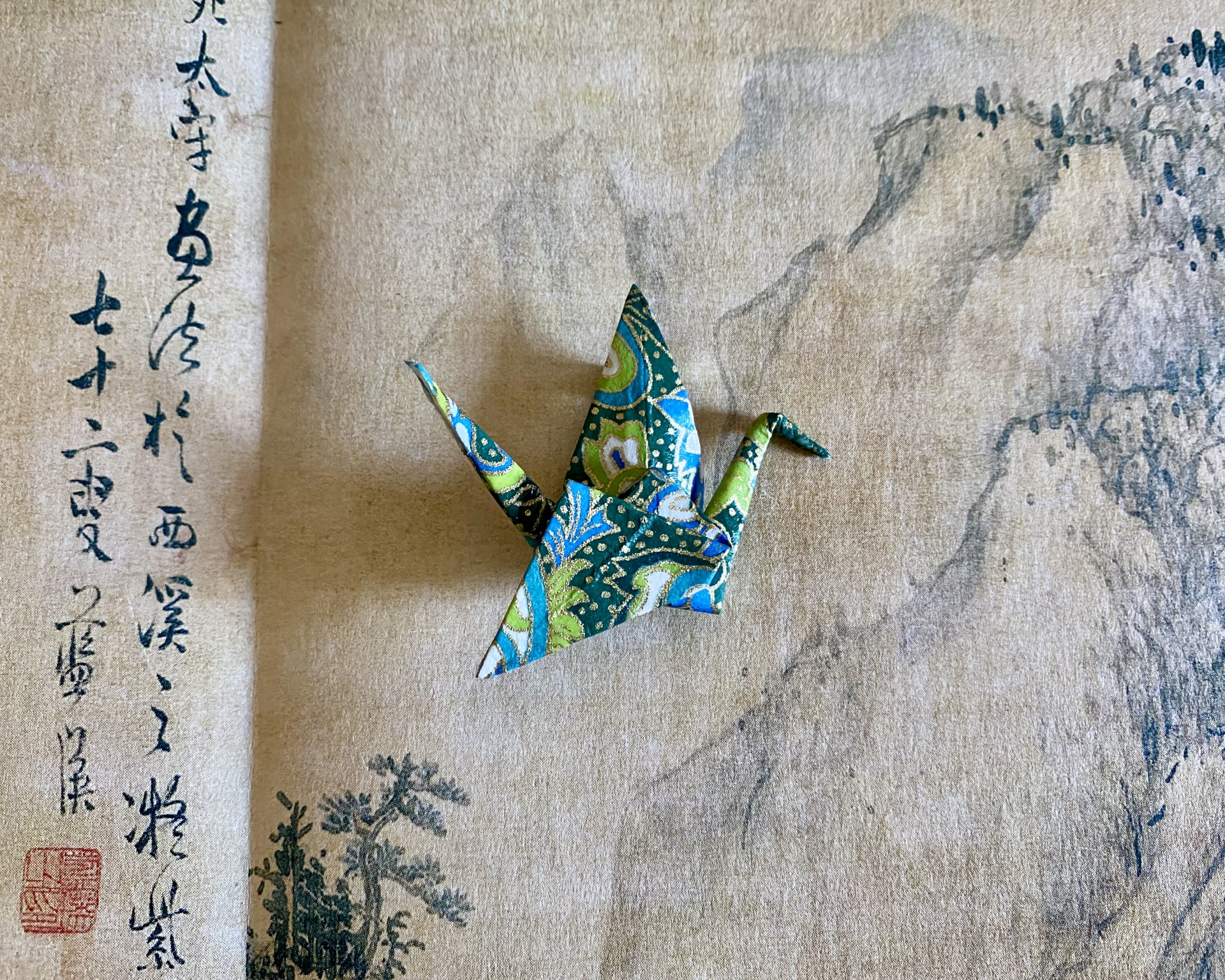 Pregadeira Tsuru Origami em papel estampado azul, verde e dourado em forma de garça sobre desenho e texto asiático.
