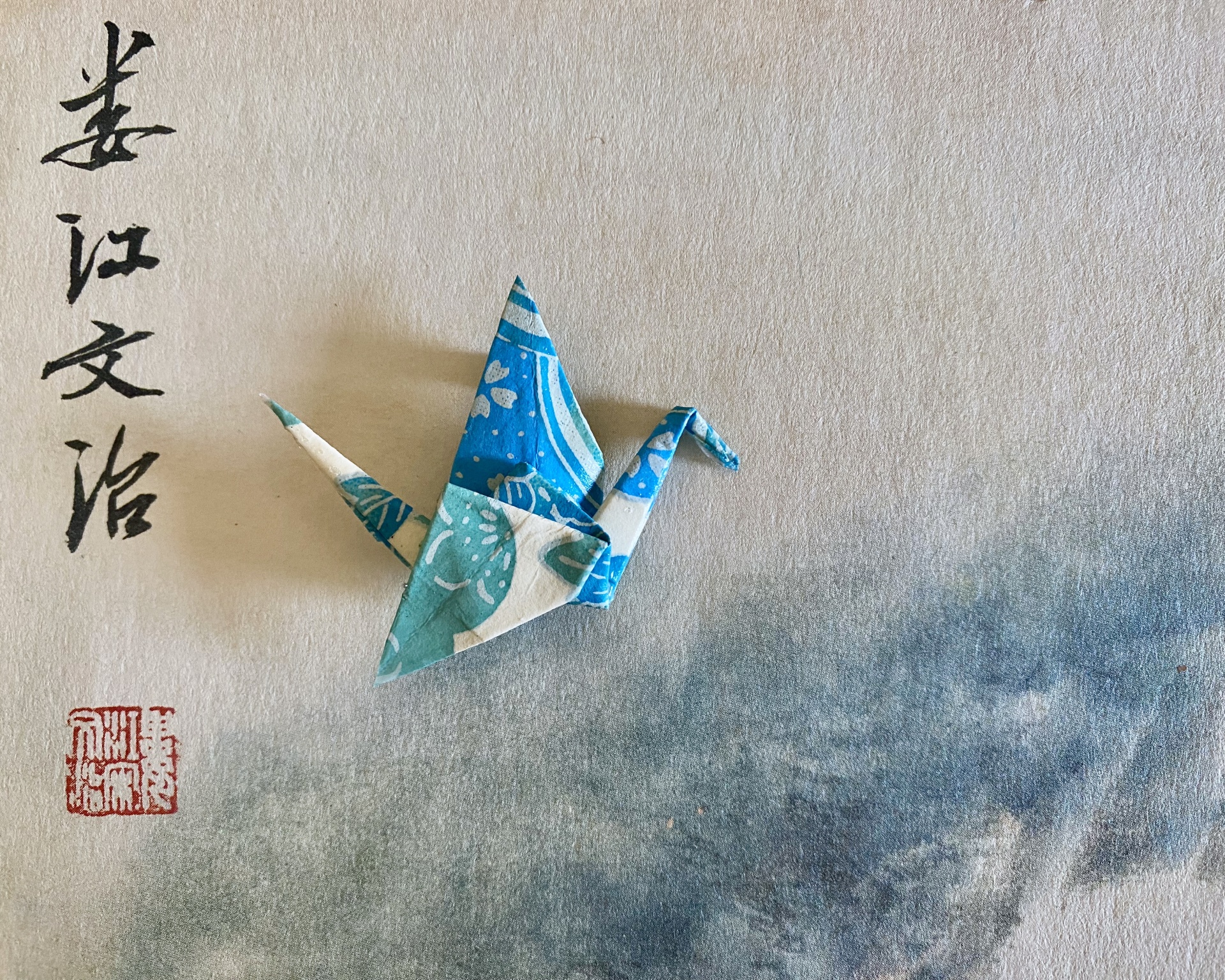 Pregadeira Tsuru Origami de grua azul e branca sobre papel com caligrafia oriental e selo vermelho.