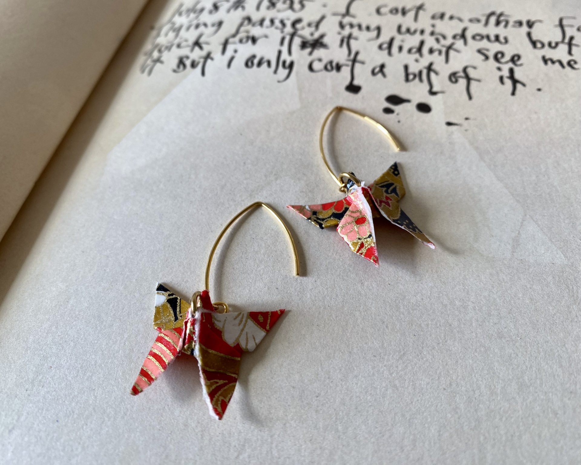 Brincos Borboleta Brincos em forma de pássaros origami coloridos e alças douradas sobre papel com escrita.