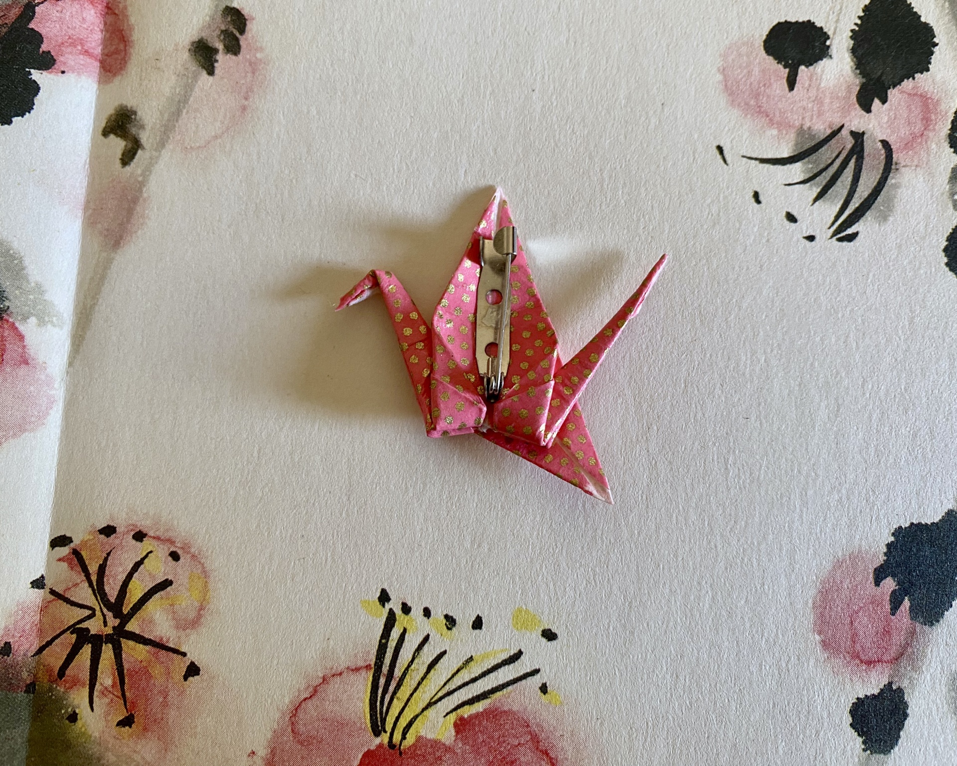 Pregadeira Tsuru Broche de origami rosa com pontos dourados e alfinete de metal sobre papel floral