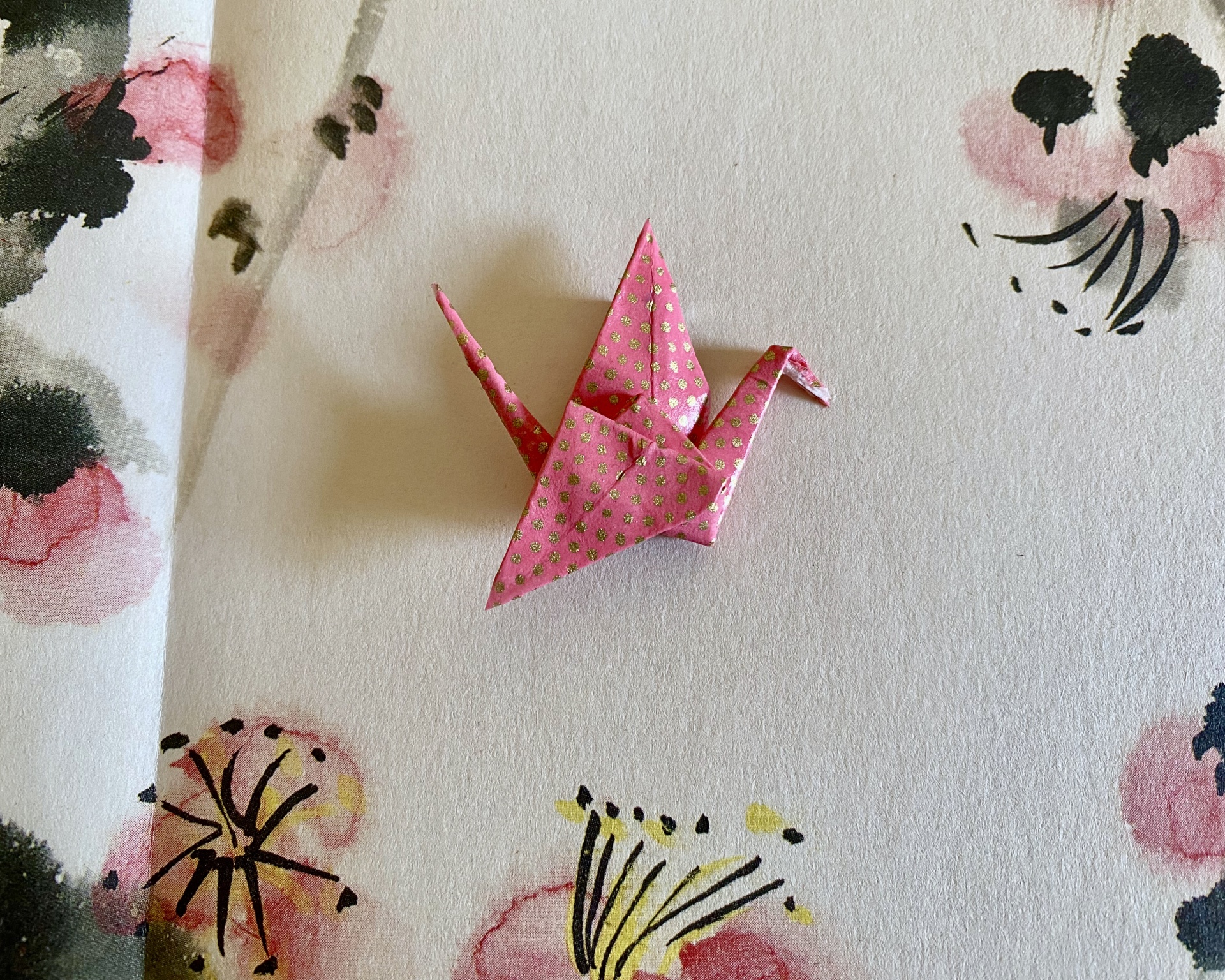 Pregadeira Tsuru Origami de pássaro rosa com padrão dourado sobre fundo branco com flores pintadas.