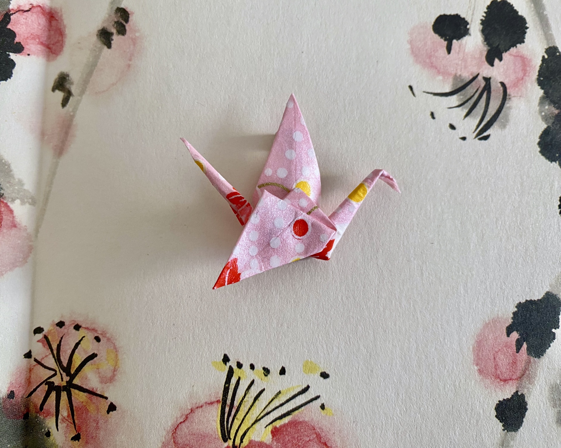 Pregadeira Tsuru Origami de pássaro em papel rosa com bolinhas e pontos coloridos sobre fundo branco floral