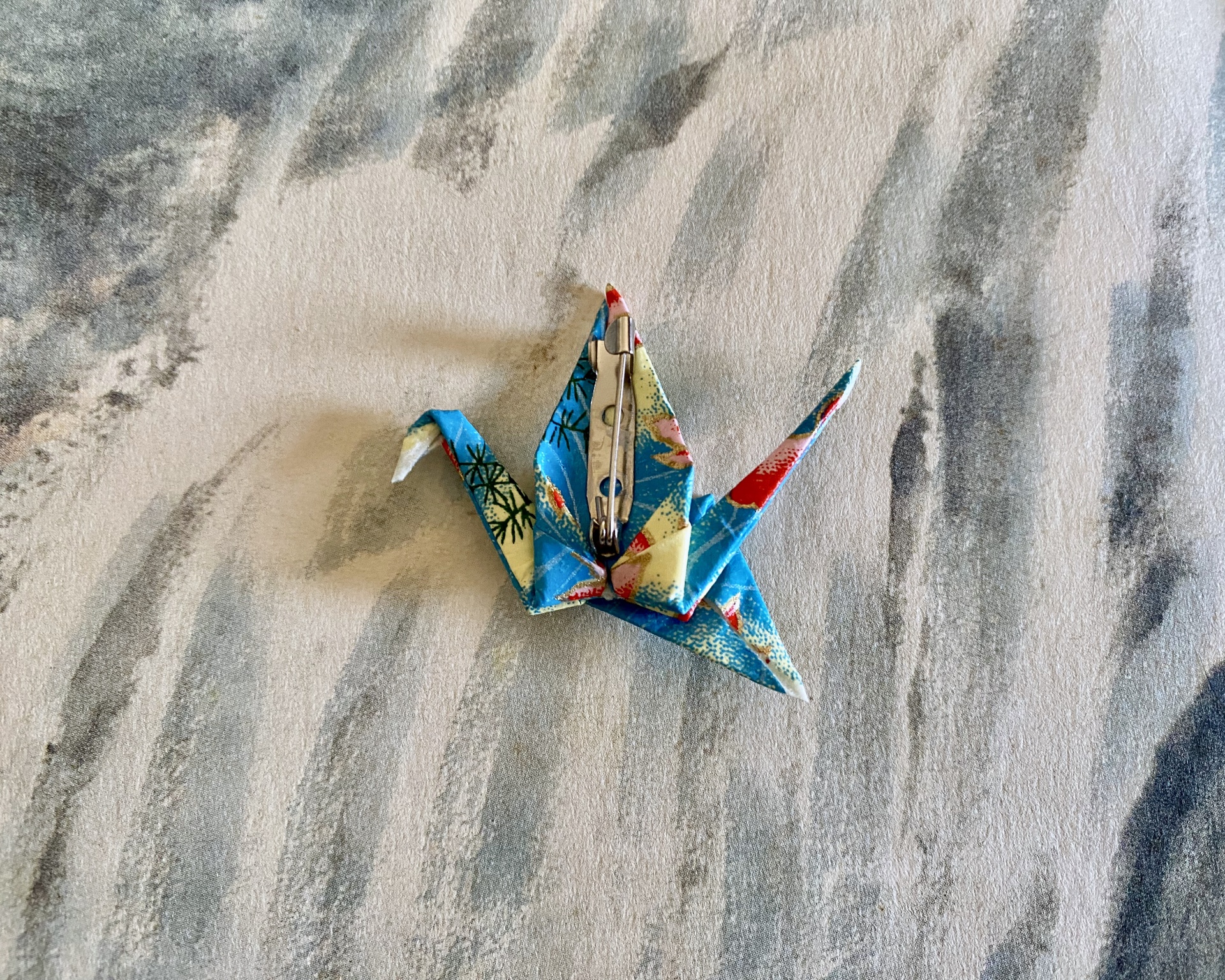Pregadeira Tsuru Broche pássaro origami de papel azul com mola metálica numa superfície de tecido cinzento.