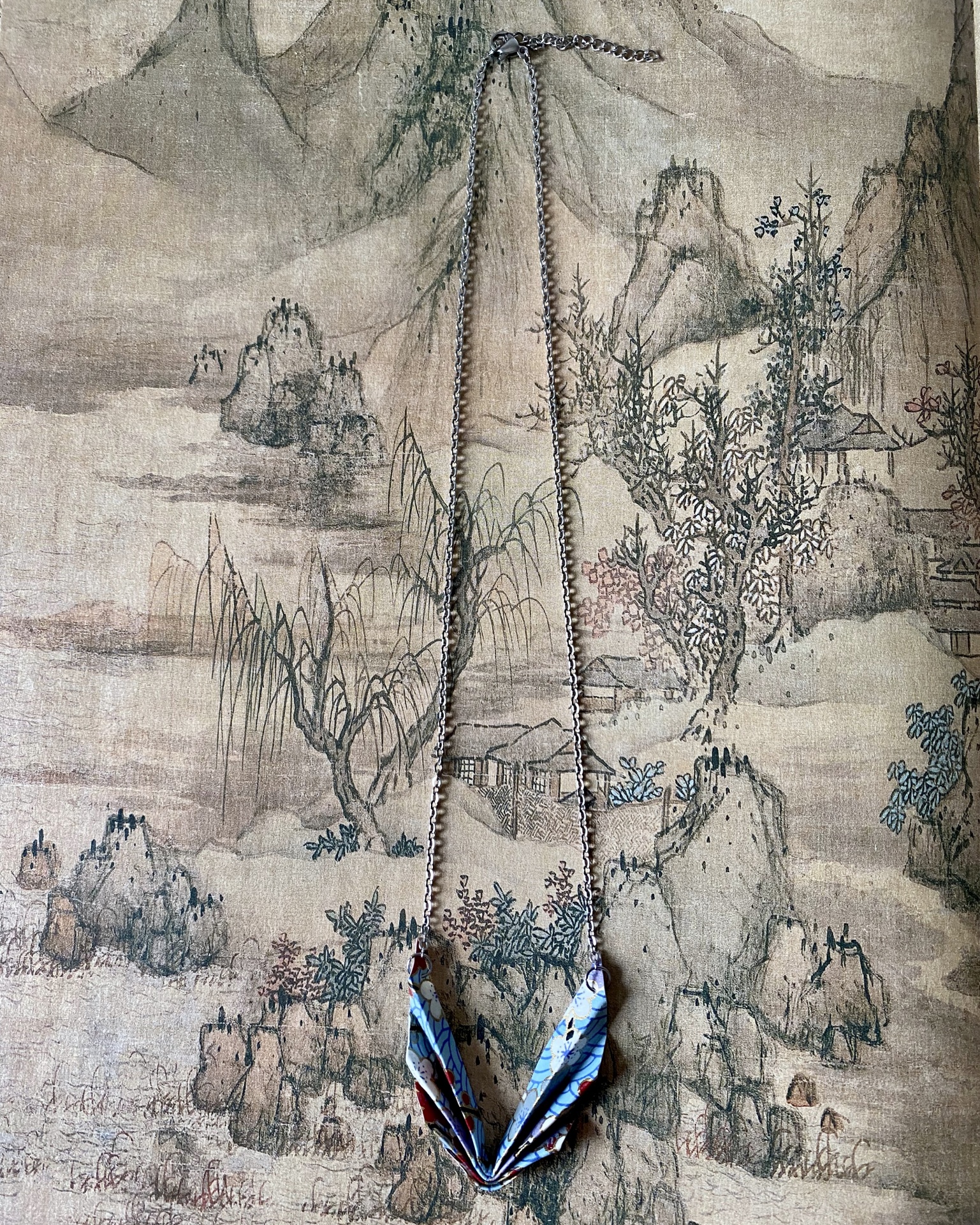 Colar Niki Colar com pendente de origami colorido pendurado num fundo com pintura de paisagem tradicional.