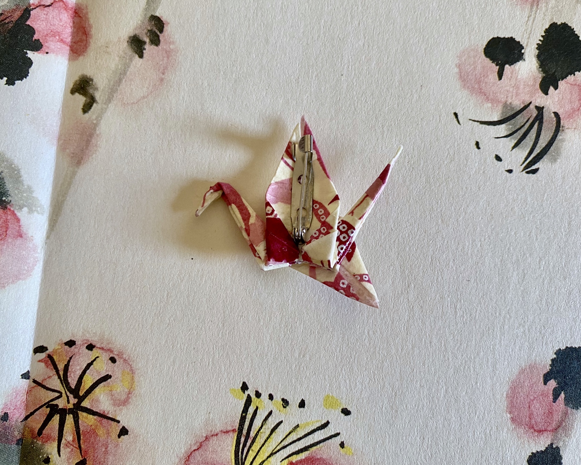 Pregadeira Tsuru Broche tsuru de origami vermelho e branco com padrão floral sobre papel com padrão de flores.