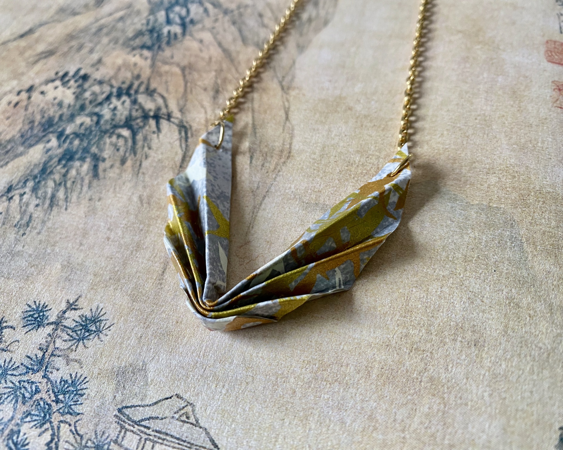 Colar Niki Colar com pendente origami colorido pendurado em corrente dourada sobre tecido bege com padrão azul